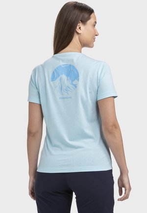 Frau mit langen braunen Haaren, die ein hellblaues T-Shirt mit einem blauen Berg und dem Schriftzug "Energizing" auf der Rückseite trägt, kombiniert mit einer dunklen Hose.