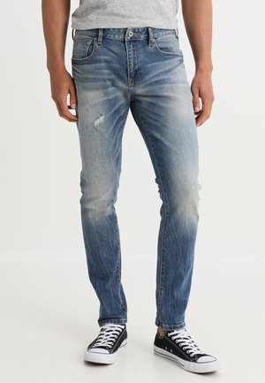 Man wearing faded blue slim-fit jeans with slight distressing and black low-top sneakers with white laces and soles.