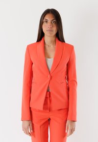 Blazer arancione su misura con rever a punta, chiusura a un bottone e due tasche frontali, abbinato a pantaloni su misura coordinati.