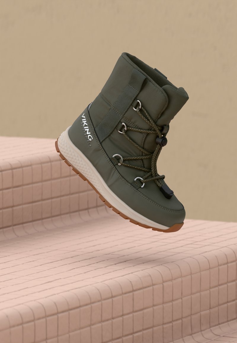 Botín de invierno aislante verde con suela blanca y cordones ajustables negros flotando sobre escaleras de cerámica beige, visible la marca "Viking" en el costado.