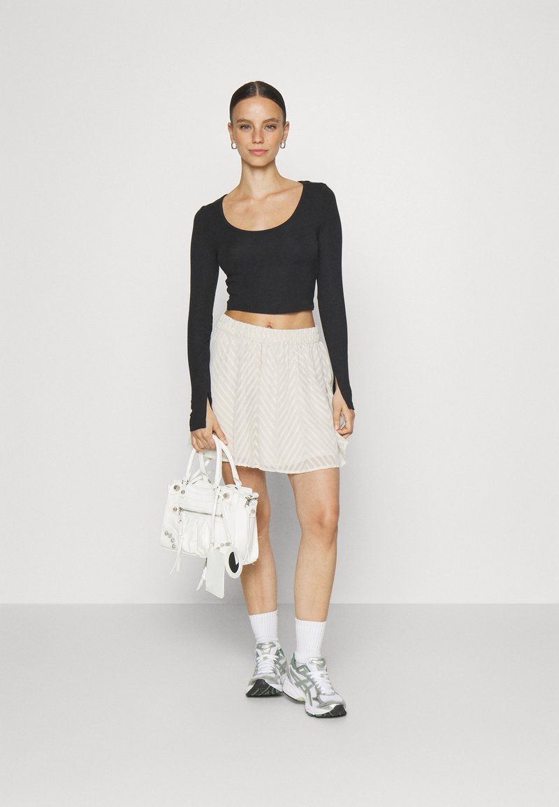 Juodas ilgomis rankovėmis crop top'as, derinamas su šviesiai BEIGE klostuotu sijonu su zigzaginiu raštu; modelis laiko mažą baltą rankinę.