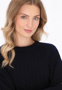 Maglione navy con lavorazione a treccia, scollo rotondo, texture morbida e vestibilità rilassata. Capelli ondulati leggeri e trucco naturale su uno sfondo neutro.