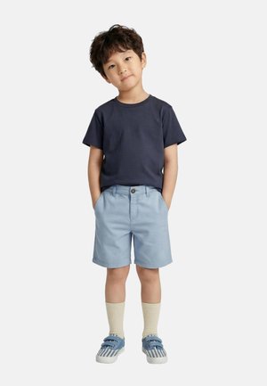 Jeune garçon debout les mains dans les poches de son short bleu clair, portant un t-shirt marine, des chaussettes crème et des baskets à rayures bleues.