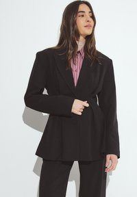 Črn eleganten blazer z oblikovanimi rameni, dvema gumboma in rahlo razširjeno silhueto, v kombinaciji z rožnato črtasto srajco spodaj.