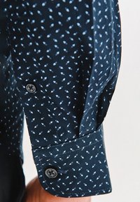 Granatowa koszula z rękawem w mały biały wzór liści, z pojedynczym guzikiem na mankiecie, noszona przez osobę na jasnym tle.