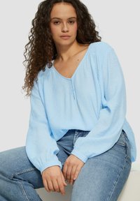 Lichtblauwe blouse met lange mouwen en V-hals, met geplooide schouders en knoopdetails bij de hals, gecombineerd met lichtblauwe denim jeans.