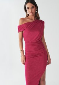 Haljina s pognutim ramenima, nabrana, od tamnoroze tkanine, s bočnim prorezom i glatkom teksturom. Naglašena nakitom na ruci.