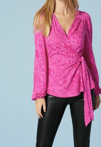 Blusa envolvente de patrón floral rosa con mangas largas y detalles de lazo, hecha de una tela texturizada. Combinada con pantalones ceñidos negros.