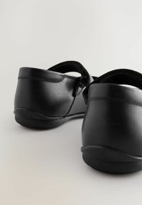 Chaussures noires en cuir avec un bout arrondi, sangle réglable et texture lisse. Elles disposent d'une semelle en caoutchouc flexible et basse avec un détail de couture subtil.