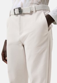 Bílý tkaný pásek s texturovaným povrchem a stříbrnou přezkou, doplněný o světle zbarvené kalhoty s hladkou látkou a bočními kapsami.