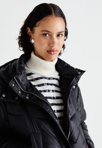 Schwarze gepolsterte Jacke mit hohem Kragen, ausgestattet mit einem Frontreißverschluss und goldenen Akzenten, über einem weißen und marine-blauen gestreiften Pullover mit Pailletten.