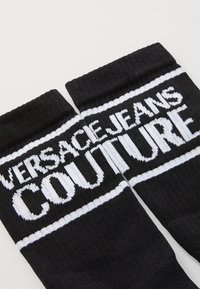 Juodos kojinės su balta užrašu "VERSACE JEANS COUTURE", su horizontaliomis juostelėmis ir tekstūruotu mezgimo dizainu.