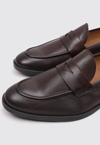Mocasines de cuero marrón con un acabado suave, punta redondeada y banda decorativa a lo largo del empeine, montados sobre una suela de goma negra.