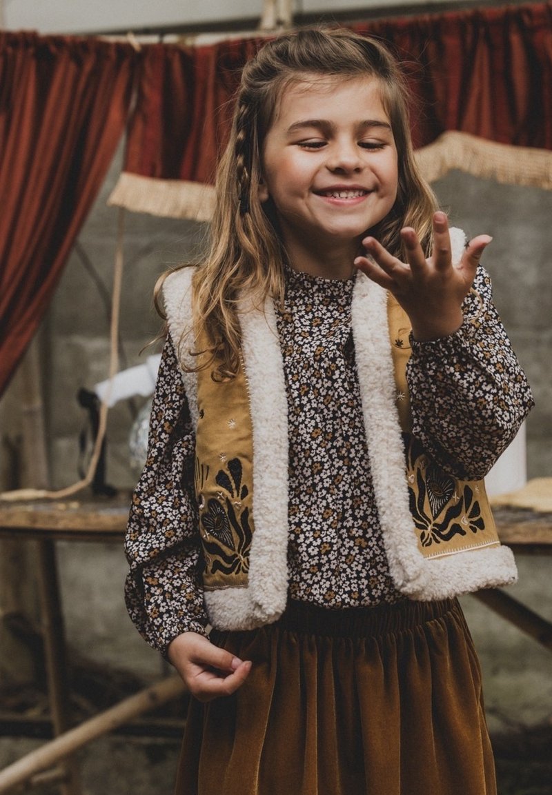 Bruine bloemenprint lange mouwen blouse, beige met schapenvacht gevoerd vest met borduursels, en een fluwelen roestkleurige rok. Het kind glimlacht terwijl ze haar hand laat zien.