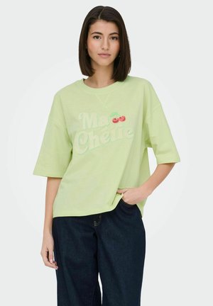 Kvinde med glat mørkt hår bærer en lysegrøn oversized T-shirt med teksten "Ma Chérie" og kirsebærgrafik, kombineret med mørkeblå jeans.