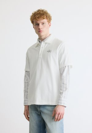 Noor mees lokkis punaste juustega, kandes valget Calvin Kleini polo särki triibulise pikavarrukaga särgi ja helesiniste teksapükstega, seisab neutraalselt.