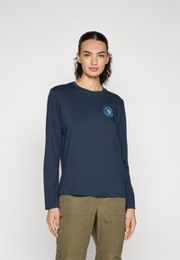 Marineblauwe longsleeve t-shirt van zachte stof met een ronde hals en een cirkelvormig logo in het blauw op de borst. Gecombineerd met olijfgroene cargobroek.