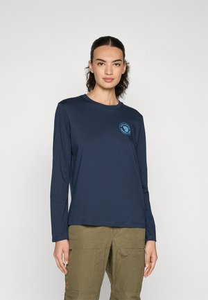 Navy long-sleeve t-shirt in soft fabric with a round neckline and a circular logo in blue on the chest. Paired with olive green cargo pants.