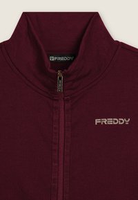 Veste zippée marron en tissu lisse, dotée d'un col haut, d'une fermeture éclair dorée et d'un logo "FREDDY" argenté sur la poitrine.