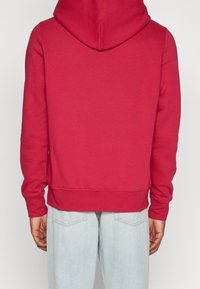 Persona vestida con una sudadera roja simple y jeans azules claros, vista desde atrás contra un fondo claro sencillo.