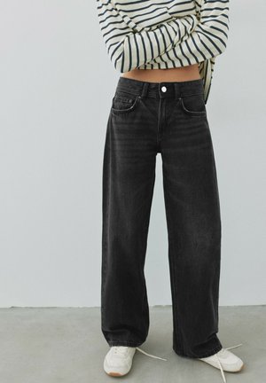 Personne portant un jean noir à jambes larges, des baskets blanches et un haut rayé à manches longues court, les bras croisés.