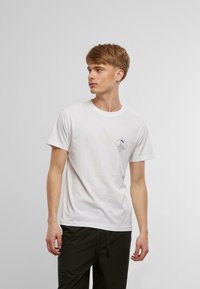 T-shirt en coton blanc à manches courtes, avec un petit graphisme d'un palmier bleu en forme de diamant sur le côté gauche de la poitrine. Col rond classique.