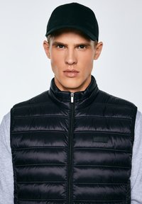 Jonge man met een zwarte baseballpet en een zwart doorgestikt mouwloos jack over een grijs shirt met lange mouwen, kijkt recht vooruit met een neutrale uitdrukking.