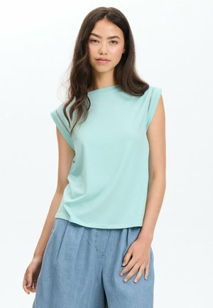Mujer joven con pelo largo y oscuro, vestida con un top sin mangas azul claro y pantalones azules holgados, de pie frente a un fondo blanco sencillo.