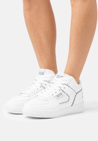 White sneakers made of smooth leather featuring perforated details, paired with silver accents and branding on the tongue and sides.