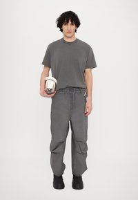 Grå bomull T-skjorte og matchende cargosbukser med elastisk midje. Modellen holder en hvit pose. Teksturert stoff med synlige sømmer. Svarte støvler.