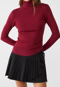 Bordeauxfärgad polotröja i mjukt strickmaterial med långa ärmar, tillsammans med en svart plisserad minikjol med en knytdetalj.