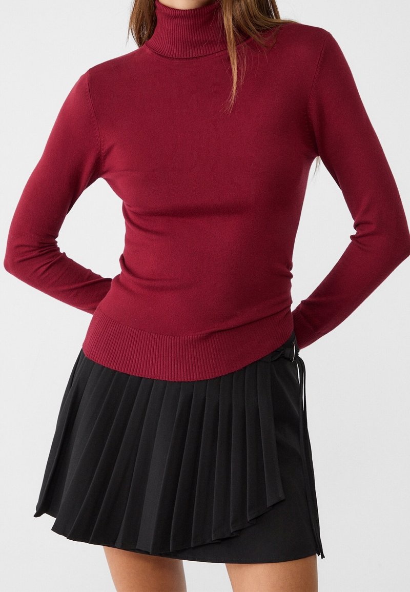 Bordeauxfärgad polotröja i mjukt strickmaterial med långa ärmar, tillsammans med en svart plisserad minikjol med en knytdetalj.