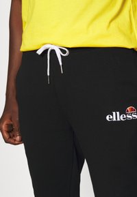 Sorte sweatpants med hvid snøre og metalspidser; har broderet "ellesse" logo i hvid med et farverigt emblem ovenfor.