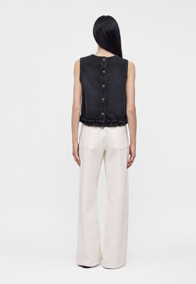 Femme aux longs cheveux foncés portant un haut noir sans manches boutoné et un pantalon blanc à jambes larges, debout, dos à l'appareil, sur un fond uni.