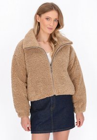 Giacca in pile marrone con esterno testurizzato, colletto alto, chiusura con zip e polsini elasticizzati. Indossata sopra una maglietta bianca e una gonna di jeans.