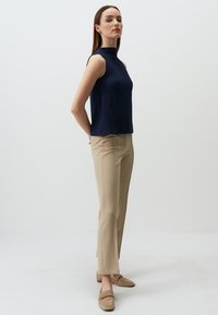 Top senza maniche blu navy in tessuto texturizzato, collo alto; abbinato a pantaloni beige su misura e scarpe slip-on marrone chiaro.