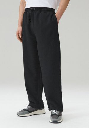 Persona che indossa pantaloni da tuta neri a vestibilità ampia con vita elastica e scarpe da ginnastica grigie, in piedi davanti a uno sfondo semplice.