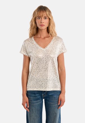 Donna con capelli biondi che indossa una maglia a maniche corte con scollo a V, bianca con stampa leopardata beige, e jeans blu, in piedi davanti a uno sfondo semplice.