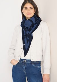 Marineblåt trekantet tørklæde med abstrakt mønster, lavet af blødt stof. Båret over en lys grå jakke, kombineret med blå jeans.