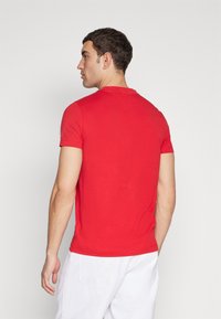 T-shirt rosso a maniche corte con collo rotondo e una texture di cotone liscia. L'orlo laterale presenta un piccolo logo del marchio. Pantaloni corti bianchi visibili.