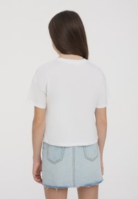 T-shirt bianco a maniche corte e a busto corto, vista posteriore, abbinato a una gonna mini in denim azzurro chiaro con orlo sfrangiato e due tasche posteriori.