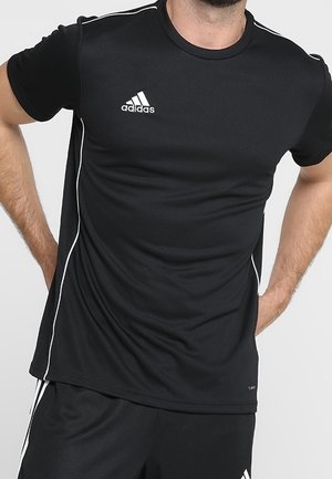 Mężczyzna w czarnej sportowej koszulce Adidas z białymi lamówkami i logo, stojący z rękami na biodrach na neutralnym tle.
