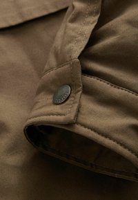 Poignet de chemise en tissu brun avec un bouton en métal visible gravé de "EST. 1982" et des coutures détaillées le long des bords.