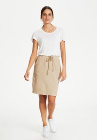 Lätt vit t-shirt i kombination med en beige kjol med dragsko i midjan. Modellen bär vita sneakers och står framför en enfärgad bakgrund.