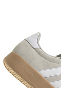 Zapatillas de ante gris claro con acentos blancos, puntera redonda y suela de goma texturizada color goma. Presenta tres rayas blancas y un cuello acolchado.