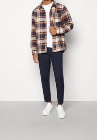 Man i marinblå byxor, vita sneakers, vit t-shirt och en uppknäppt rutig skjorta i orange, marin och krämfärgat, står på en grå golv.