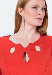 Rotes Kleid mit einem runden Ausschnitt und zwei Aussern. Weicher Stoff, figurbetonte Silhouette, ergänzt durch hängende silberne Ohrringe.