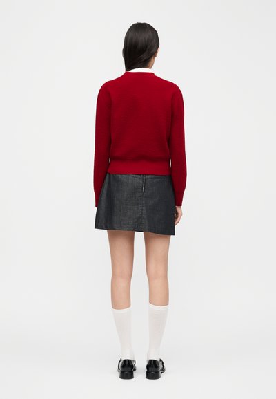 Femme aux cheveux foncés portant un pull rouge à manches longues, une jupe en denim noire, des chaussettes blanches montant jusqu'aux genoux et des chaussures noires, debout de dos.