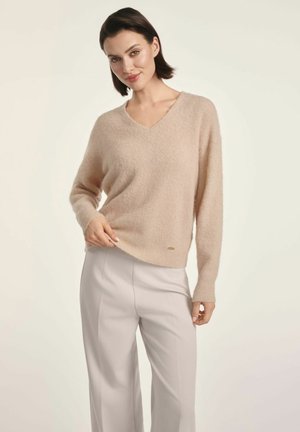 Beżowy, puszysty sweter w serek z długimi rękawami, delikatnie dopasowany. Noszony z jasnymi, wysokim stanem spodniami. Małe logo na dolnej krawędzi.