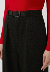 Zwarte stoffen broek met een gestructureerde afwerking, voorzien van een riem met een zilveren gesp. Gecombineerd met een rode longsleeve shirt.